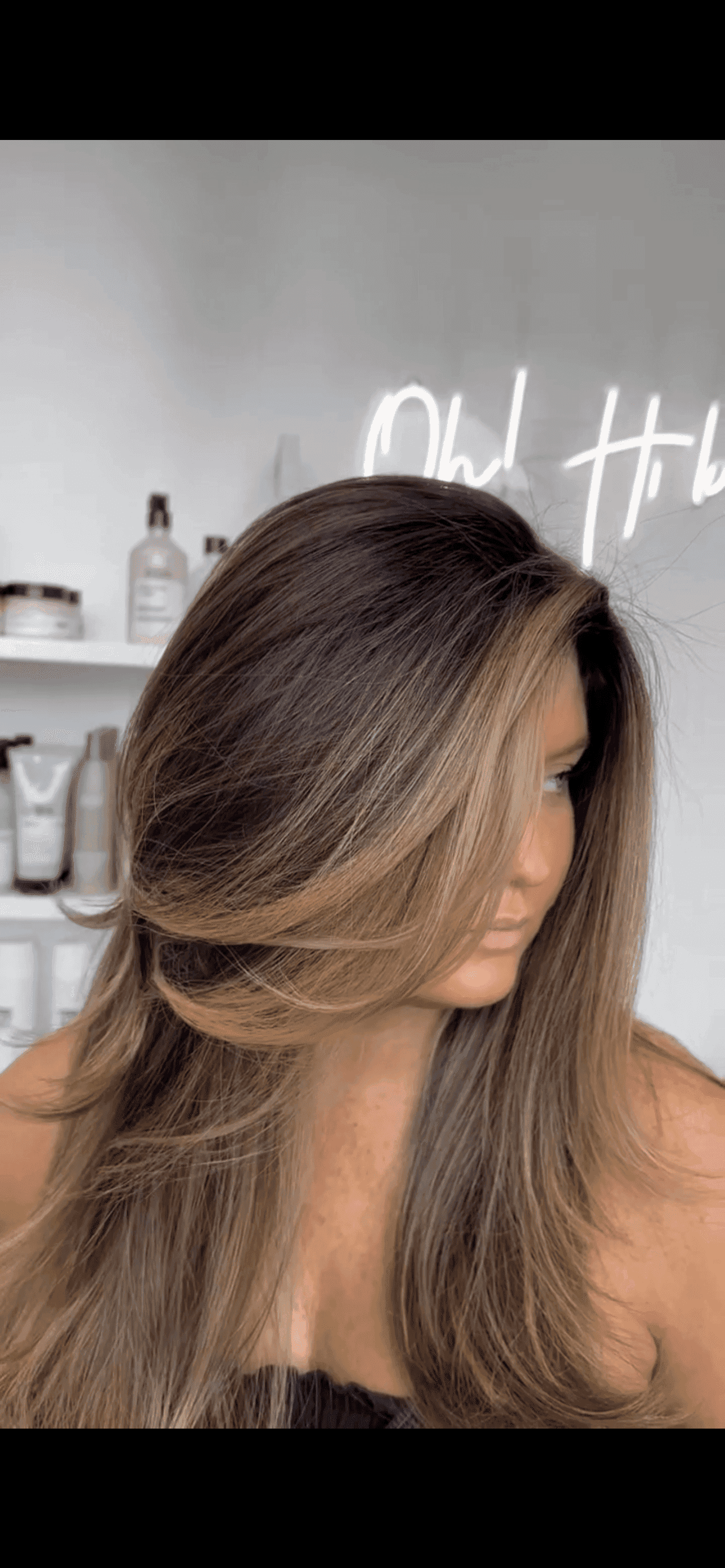 Woman with voluminous layered hair, blonde highlights, in a salon with hair products on shelves.